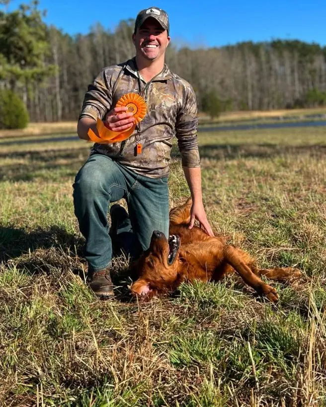 Matthew kneeling in a field with an orange ribbon rosette, his red golden retriever rolling beside him.